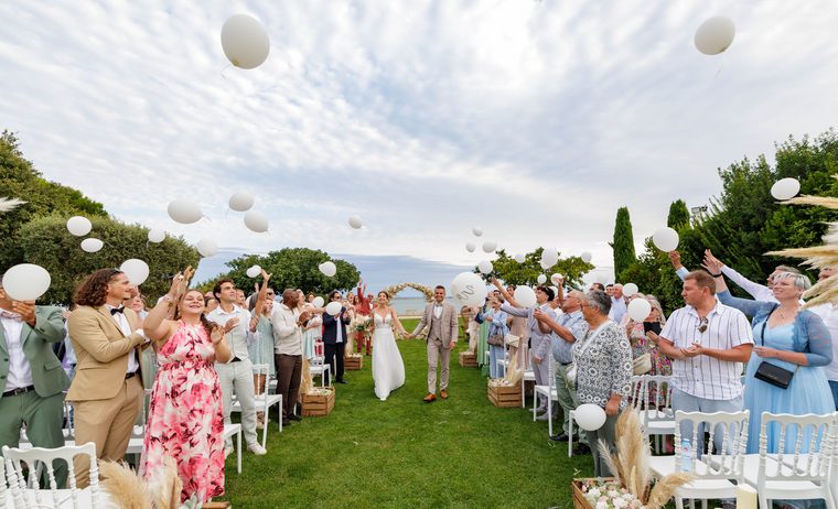 Les mariés remontent l’allée main dans la main à la sortie de la cérémonie, sous les ballons blancs lâchés par leurs invités debout